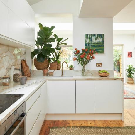 vitt kök med bänkskivor i neutral marmor, vita vägg- och basenheter, konstverk, trägolv med matta, växter