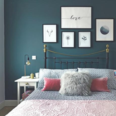 Tumši zila siena aiz gultas ar pelēku raksta gultas veļu un rozā spilveniem ar metāla rāmi un apdrukām uz sienas