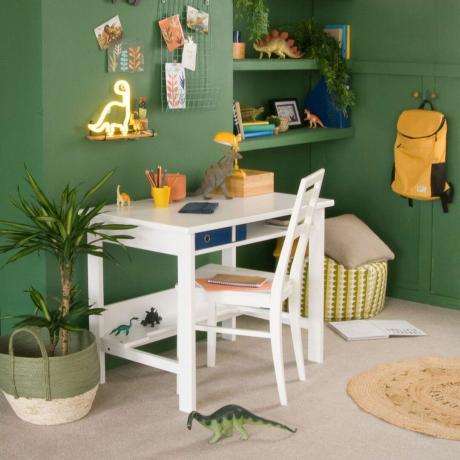 Окрашенная в зеленый цвет детская спальня с белым письменным столом и письменным стулом, а по комнате разбросаны динозавры.