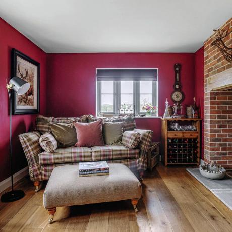 غرفة معيشة صغيرة بجدران حمراء ومدفأة من الطوب وأريكة مدمجة ذات مقعدين في قماش مربعات
