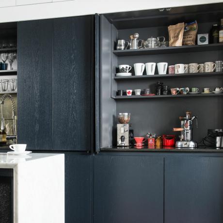 Mörkgrå köksskåp med dubbla dörrar öppna för att visa hyllor fulla av muggar och en bänkskiva kaffestation inställd