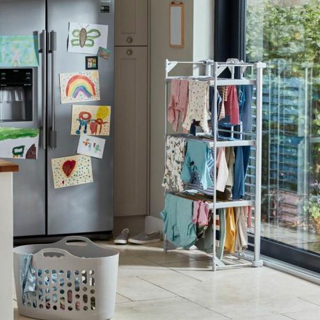 apsildāms drēbju ventilators, pilns ar drēbēm, kas atrodas virtuvē blakus lielajam ledusskapim un logam