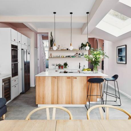 O bucătărie-lounge modernă cu vopsea roz pentru pereți, trio de corpuri de iluminat cu pandantiv alb și insulă din lemn, dulapuri albe încorporate și aparat de cuptor