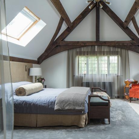 Спальня на мансарді з дерев’яними балками та вуалевими шторами від Розелінд Вілсон