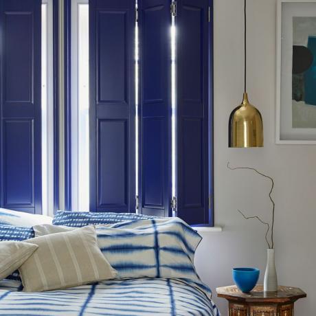 kamar tidur dengan sghutters biru solid di jendela dan tempat tidur di depan ditutupi sprei biru dan putih cetak ikat