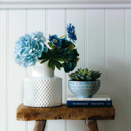 Сини цветя в бяла ваза върху дървена табуретка, бяла ламперия, боядисана в синьо стена