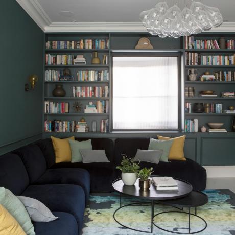 Et hyggeligt stueområde med indbygget bogreol, der viser en række forskellige bøger, mørkeblå L-formet sektionssofa og blågult områdetæppe med abstrakt motiv og statement-lysekronelys armatur