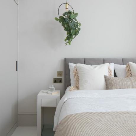 Sypialnia z białymi ścianami, neutralnym białym i szarym łóżkiem i wiszącą rośliną
