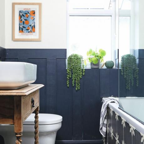 Dunkles, wandgetäfeltes Badezimmer mit dekorativem Bodenbelag, hängender Wandkunst und Pflanzen