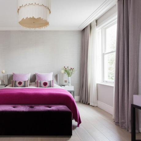 camera da letto con carta da parati grigia testurizzata, pavimento in legno in stile calce, pouf rosa brillante e plaid, cuscini e tende lilla, paralume importante, comodini coordinati