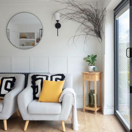 Vitt vardagsrum med panelväggar, grå fåtöljer och rund spegel