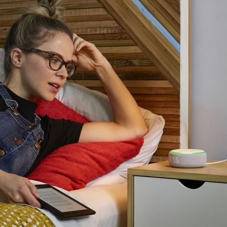 dívka ležící v posteli sedí a mluví s chytrým reproduktorem na jejím nočním stolku