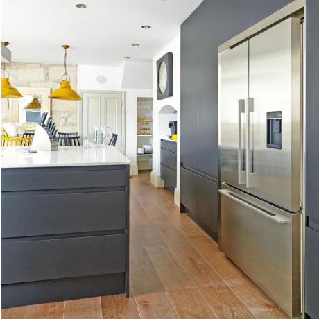 virtuvė su medinėmis grindimis, garstyčių pakabinami šviestuvai, tamsiai pilkos spalvos spintelės ir sidabrinis šaldytuvas-šaldiklis