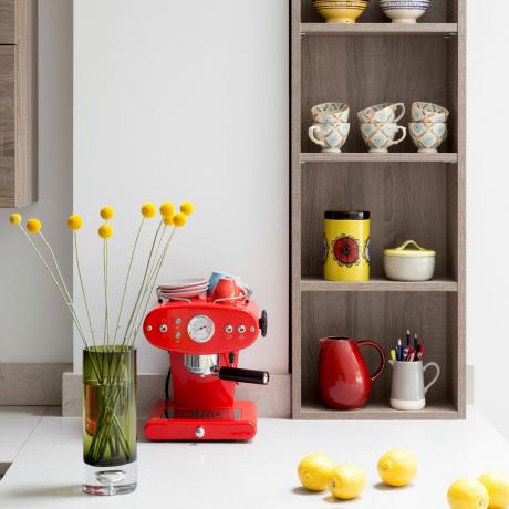 Balta virtuvė su medinėmis lentynomis, užpildytomis puodeliais, ir raudonu kavos aparatu ant balto stalviršio