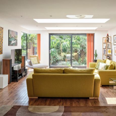 pomysły na podłogę drewnianą w salonie, pokój dzienny z podłogą z twardego drewna, dywan, dwie żółte sofy, krzesło, poduszka w stylu retro, pomarańczowe zasłony, widok na ogród, telewizor, moduł multimedialny