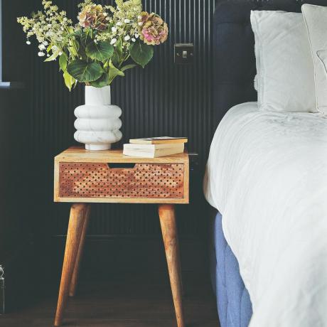 Un dormitor cu un pat cu cearșafuri albe și o noptieră din lemn cu o vază cu flori