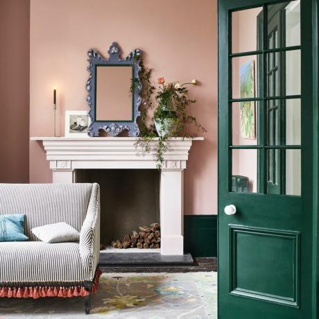 ідеї кольору плінтуса, рожева вітальня з темно-зеленими дерев’яними виробами, кольоровий плінтус, вінтажний килим, сіре дзеркало, смугастий диван