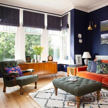 turuncu kanepe, adaçayı yeşili koltuk ve tabure, desenli kilim, yüzyıl ortası büfe, grafik panjurlar, duvar lambası, cesur grafik minderler içeren mavi oturma odası