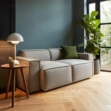 Swyft pilka sofa su šoniniu stalu ir lempa, medinėmis grindimis ir augalu dešinėje
