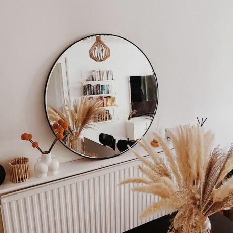 Dunelm apaļais spogulis Annijas mājās uz radiatora plaukta, atspoguļojot grāmatu skapi un televizoru ar pampas zāli un svecēm