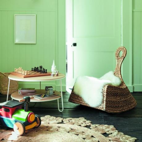 Gröna väggar, trägolv, texturerad matta, träbord med schackbräde och liten rottingstol med fluffiga plagg