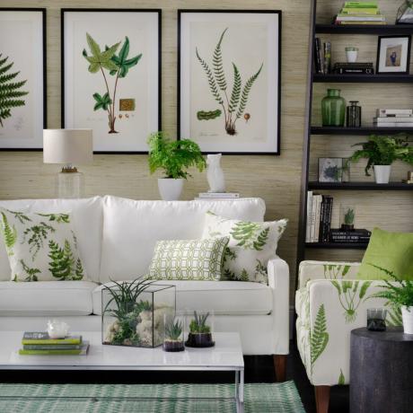 غرفة معيشة نباتية مع أريكة بيضاء وكرسي بذراعين بطباعة نباتية ومطبوعات نباتية على الجدران وطاولة قهوة منخفضة بيضاء وعصارة ووحدة رفوف مع كتب ومزهريات ونباتات