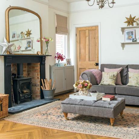 Белая гостиная с серым диваном и подставкой для ног, открытый кирпичный камин с дровяной горелкой, зеркало над камином