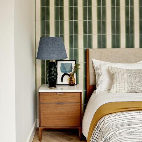 Luxusná spálňa s dekoráciou krémových a zelených dlaždíc, posteľ, džínsovo modré tienidlo, drevený a biely nočný stolík a umenie v čiernom ráme
