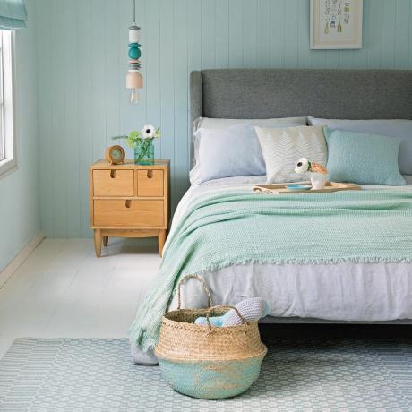 Šviesiai mėlynas miegamasis su pilka minkšta lova ir švelnių pastelinių atspalvių patalyne