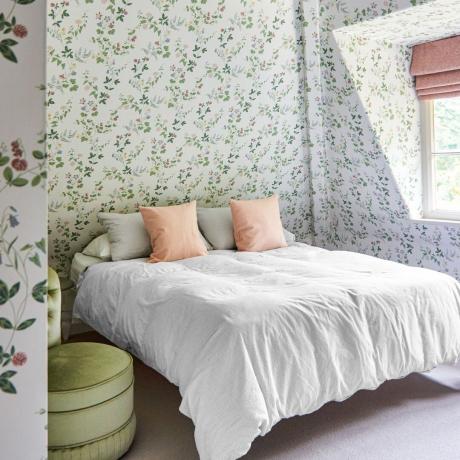 Dormitor cu tapet floral pe pereți și tavan cu husă de pilota albă și perne roz și jaluză romană