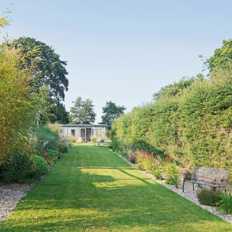 Suuri puutarha, jossa pitkä vihreä hoidettu nurmi, puutarhapenkki, sorakoristelu molemmilla puolilla ja laivastonsininen hytti suunnitelman takana