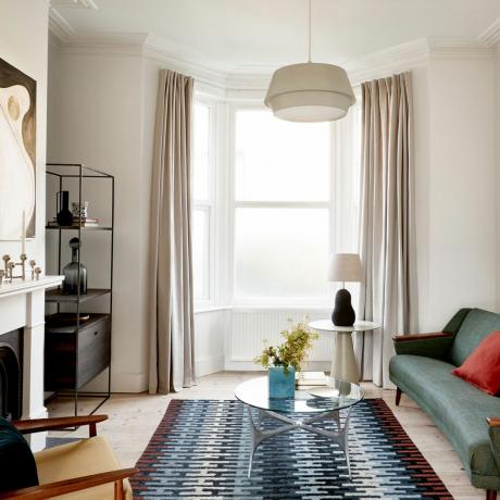 Sala de estar branca com janela saliente, cortinas compridas, tapete estampado, sofá Scandi, mesa de centro de vidro, lareira com castiçal e obras de arte abstratas