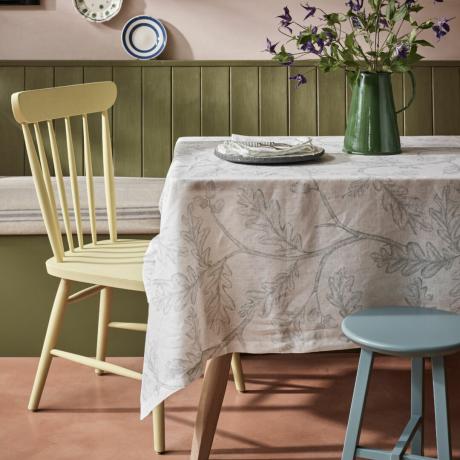 liten spisestue med sittebenk, gulmalt stol, blåmalt krakk, mønstret duk, vase med klematis, tallerkener på vegg, malt not og fjær