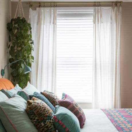 Dormitor vopsit în bej, cu pat decorat cu perne colorate și jardinieră agățată lângă fereastră mare cu jaluzele și perdele transparente