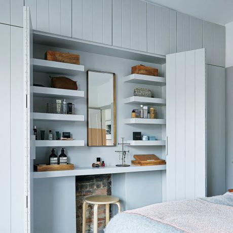 Un dormitor cu uși mari de dulap deschise pentru a arăta o masă de toaletă înăuntru, lemn vopsit în gri pal, cu perete încorporat de dulapuri și depozitare