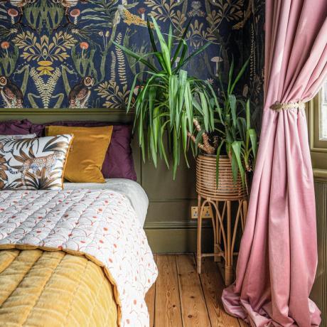 Miegamasis su tapetais akcentuota siena ir sienų apdaila, brangakmenių atspalvių patalynė ir kambarinis augalas