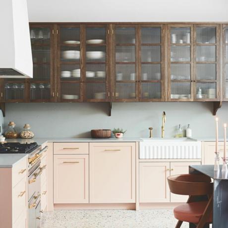 kuhinja z belo kuhinjsko napo in belimi stenami nad stenskimi enotami, bledo modre delovne plošče in hrbtna stran, pomivalno korito, bledo rožnate enote, teraco tla, miza in stol ob strani