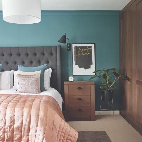 Makuuhuoneessa sänky, harmaa sängynpääty ja vaaleanpunainen sängynpääty, vieressä yöpöytä ja kasvi, jonka takana on tummansininen seinä