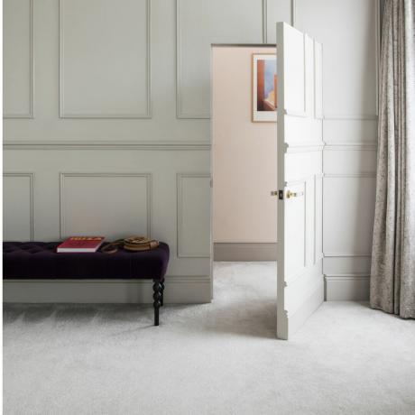 क्रीम कालीन, पैनलिंग वाली क्रीम दीवारें और खुले दरवाज़े और बैंगनी रंग के फुट स्टूल वाला कमरा