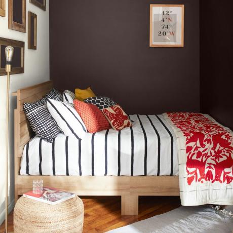 ідеї кольорів для маленької спальні, маленька спальня з шоколадно-білими стінами, дерев’яне ліжко, смугаста постільна білизна червоного кольору, подушки, твори мистецтва, табурет з ротанга, дерев’яна підлога