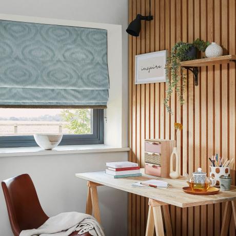 مكتب منزلي بجدران مغطاة بألواح خشبية وستارة رومانية زرقاء على النافذة