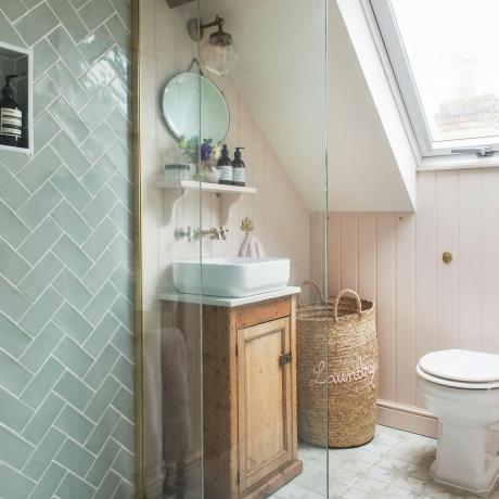 Rožinis vonios kambarys su sienų apdaila, pastelinėmis plytelėmis, perdirbta kosmetine ir praustuvu ant stalo