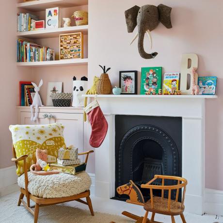 Švelniai rožinės spalvos dažytas vaikų kambarys su baltu židiniu ir dramblio galva ant sienos viršuje