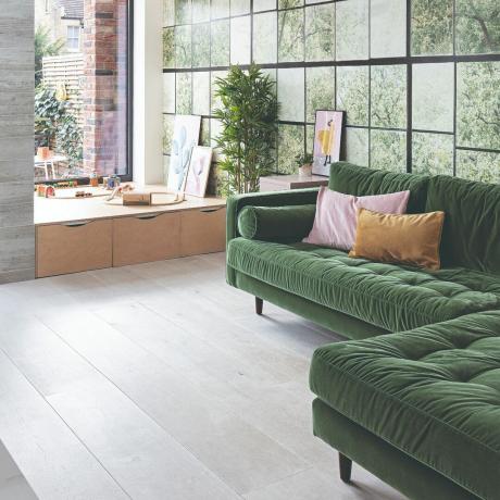 Žalia kampinė sofa svetainėje su dideliais langais