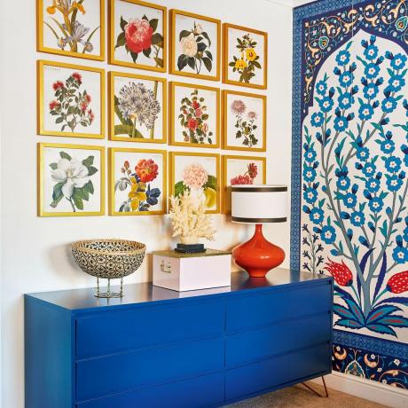 Värikäs makuuhuone, jossa sininen senkki säilytystä varten ja galleriaseinä kasvitieteellisiä taidekuvia