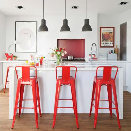 Balta virtuve ar sarkaniem bāra krēsliem un piekarināmu apgaismojumu virs salas