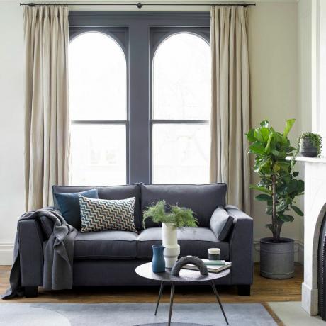 beigenvärinen olohuone, jossa harmaa antrasiitin ja harmaat verhot, koristeltu kasveilla ja harmaa sohva tyynyillä ja pyöreä teräksinen sohvapöytä