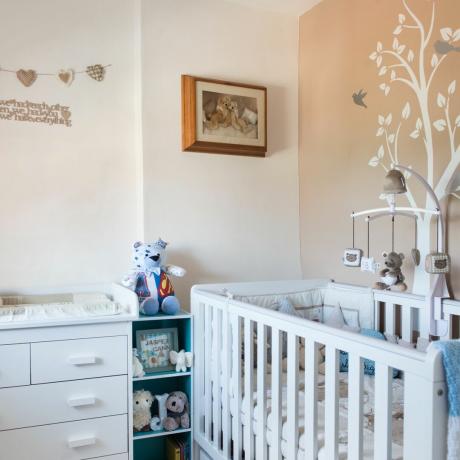 나무 프레임과 벽 화환으로 완성된 나무 디자인이 있는 베이지색 벽 앞에 있는 흰색 유아용 침대