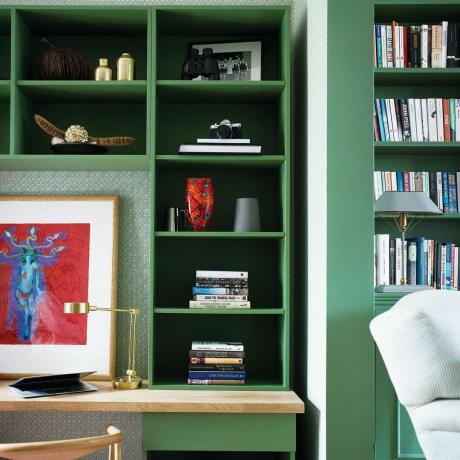 Šalia laiptelių įmontuota rašomojo stalo zona, žalia tapetuota siena ir įmontuotos žalios knygų lentynos