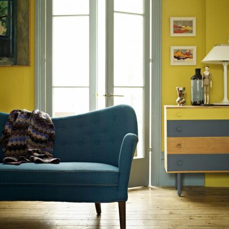 svetainės dažų spalvos 2023, geltona svetainė su pilkai ir geltonai dažyta bortu, meno kūriniai, mėlyna retro stiliaus sofa, medinės grindų lentos